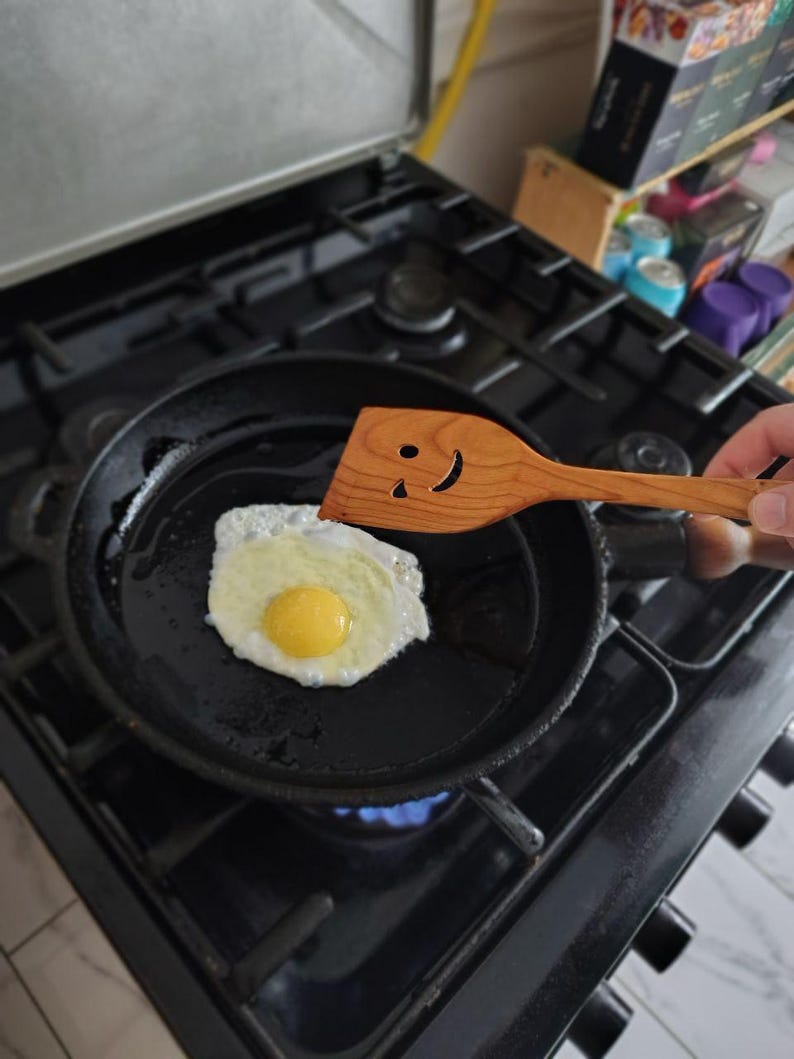 May include: A fried egg in a black frying pan on a gas hob. A wooden spatula with a smiley face is used to flip the egg. The egg has a yellow yolk and white egg whites. The hob is black.