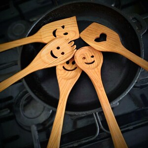 May include: A selection of wooden kitchen utensils, including spatulas and spoons, arranged around a cast iron frying pan. Each utensil features a carved smiley face. The wood is light brown in colour.