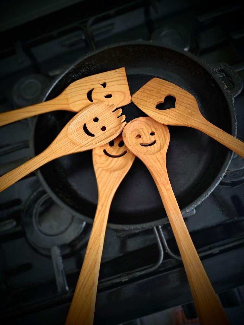 May include: A selection of wooden kitchen utensils, including spatulas and spoons, arranged around a cast iron frying pan. Each utensil features a carved smiley face. The wood is light brown in colour.