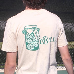 May include: Cream-colored t-shirt featuring a green graphic of pickleballs in a jar with the word "PICKLE" on a tag. The word "BALL" is printed in a script font. The shirt is made of a soft material.
