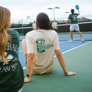 May include: Two people sitting on a blue and green tennis court, wearing t-shirts with pickleball-themed graphics. The shirts are green and tan. Other people are playing pickleball in the background.