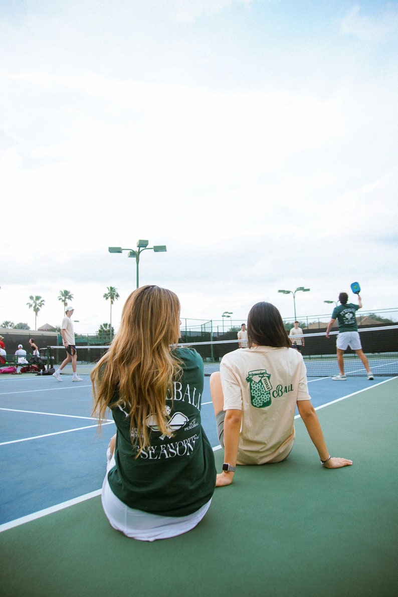 May include: Two people sitting on a blue and green tennis court, wearing t-shirts with pickleball-themed graphics. The shirts are green and tan. Other people are playing pickleball in the background.