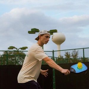 May include: A person playing pickleball on a blue court, wearing a beige t-shirt, dark shorts, and a white cap. The pickleball paddle is blue and black with a yellow ball. A water tower is in the background.