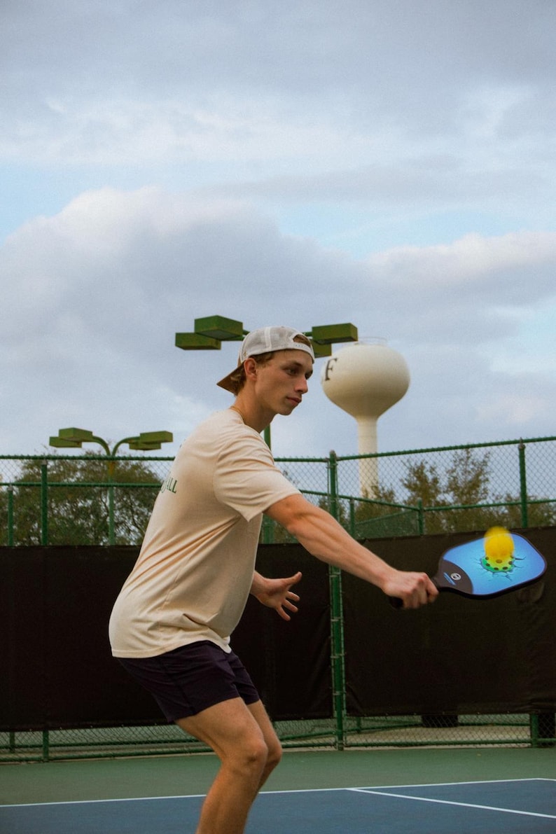 May include: A person playing pickleball on a blue court, wearing a beige t-shirt, dark shorts, and a white cap. The pickleball paddle is blue and black with a yellow ball. A water tower is in the background.