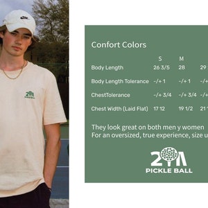 May include: A person wearing a cream-colored t-shirt with a small green pickleball logo. The shirt has a crew neck and short sleeves. The image also includes a size chart with measurements in inches and the text "21 Pickle Ball".