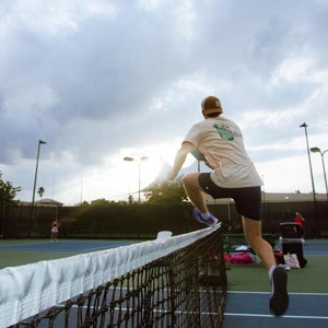 May include: A person jumping over a tennis net on a blue court. The person is wearing a tan t-shirt, dark shorts, and a brown cap. A green tennis ball is on the court. The sky is cloudy.