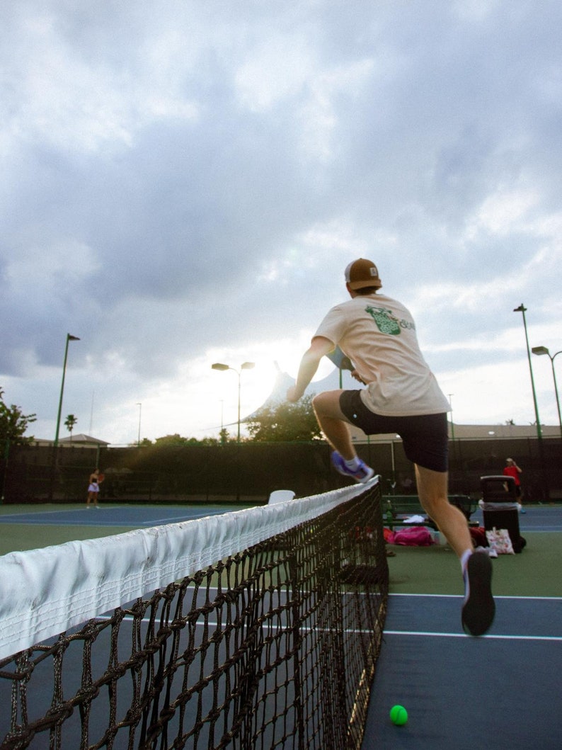 May include: A person jumping over a tennis net on a blue court. The person is wearing a tan t-shirt, dark shorts, and a brown cap. A green tennis ball is on the court. The sky is cloudy.