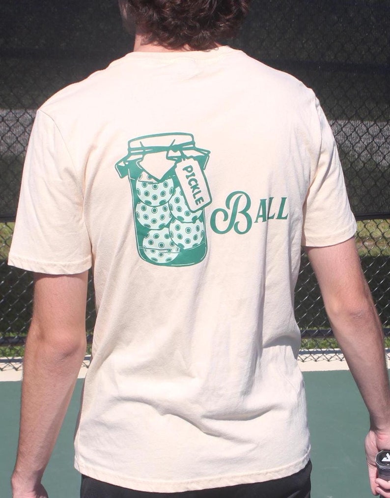 May include: Cream-colored t-shirt featuring a green graphic of pickleballs in a jar with the word "PICKLE" on a tag. The word "BALL" is printed in a script font. The shirt is made of a soft material.