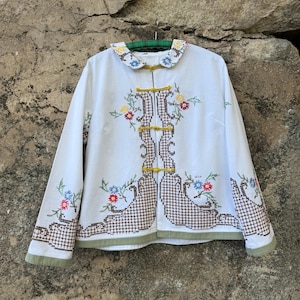 Könnte beinhalten: Eine weiße Langarmjacke mit abgerundetem Kragen und blauer Blumenstickerei, gelben Knebelverschlüssen sowie braunen Kreuzstich-Gittermustern, die auf der Vorderseite und den Ärmeln mit bunten Blumenmotiven und einem hellgrünen Saum verziert ist.