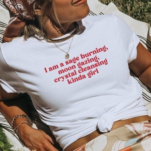 K&ouml;nnte beinhalten: Ein wei&szlig;es T-Shirt mit dem Text "I am a sage burning, moon gazing, crystal cleansing kinda girl" in roten Buchstaben. Das Shirt wird von einer Person getragen, die auf einer gestreiften Decke mit einem Buch mit dem Titel "Anne of Green Gables" und einem Blumenstrau&szlig; sitzt.