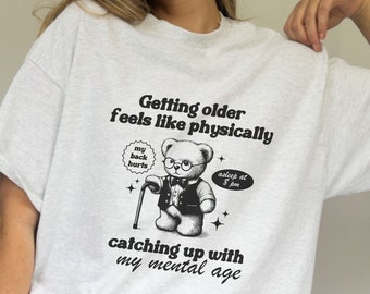 Könnte beinhalten: Ein weißes T-Shirt mit einem schwarzen Aufdruck eines Teddybären mit Brille und Anzug. Der Text auf dem Shirt lautet "Getting older feels like physically my back hurts asleep at 8 pm catching up with my mental age".