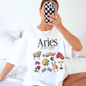 May include: White t-shirt with the text "Aries Energy" printed on the front. The shirt features a variety of graphic images including a ram, a car, boxing gloves, a disco ball, a trophy, a rose, and checkered flags.