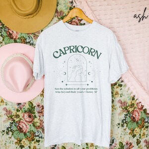 Puede incluir: Una camiseta blanca con un dise&ntilde;o gr&aacute;fico verde que dice "CAPRICORN" con una imagen estilizada de una persona con una luna creciente y estrellas. El texto debajo del gr&aacute;fico dice "has the solution to all your problems wise beyond their years bossy AF".
