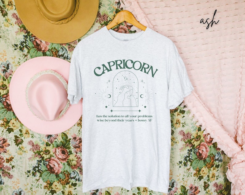 Puede incluir: Una camiseta blanca con un dise&ntilde;o gr&aacute;fico verde que dice "CAPRICORN" con una imagen estilizada de una persona con una luna creciente y estrellas. El texto debajo del gr&aacute;fico dice "has the solution to all your problems wise beyond their years bossy AF".