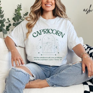 Puede incluir: Una camiseta gris con el texto "CAPRICORN" en letras verdes. La camiseta tambi&eacute;n presenta un s&iacute;mbolo astrol&oacute;gico verde y el texto "has the solution to all your problems wise beyond their years + bossy AF".