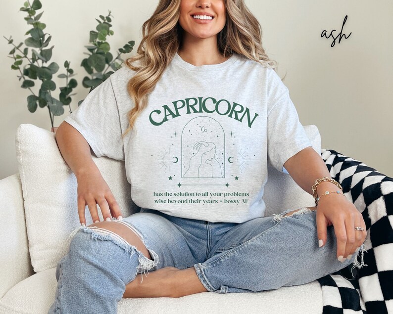 Puede incluir: Una camiseta gris con el texto "CAPRICORN" en letras verdes. La camiseta tambi&eacute;n presenta un s&iacute;mbolo astrol&oacute;gico verde y el texto "has the solution to all your problems wise beyond their years + bossy AF".