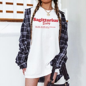 May include: A white t-shirt with the text "Sagittarius Baby" in red lettering. The shirt is worn by a person with long brown hair and a black and white checked shirt. The text on the shirt reads "Wild heart, free spirit, and a natural born storyteller. Catch me if you can."