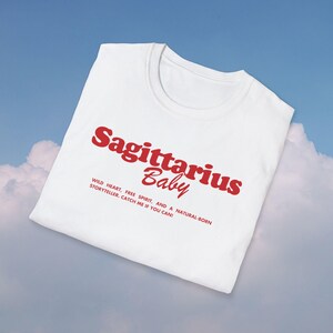 May include: White t-shirt with red text that reads "Sagittarius Baby" and a description below: "WILD HEART, FREE SPIRIT, AND A NATURAL-BORN STORYTELLER. CATCH ME IF YOU CAN!"