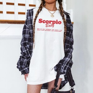 May include: A white t-shirt with a red and black graphic that says "Scorpio Baby" and the text "Sweet but spicy, mysterious, and always a step ahead. I'll keep your secrets, but I'll never forget."