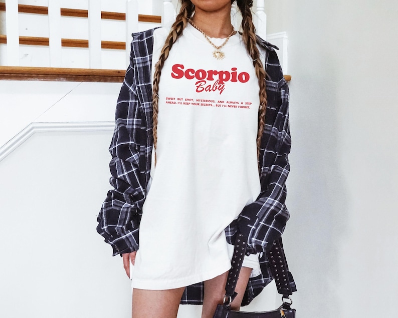 May include: A white t-shirt with a red and black graphic that says "Scorpio Baby" and the text "Sweet but spicy, mysterious, and always a step ahead. I'll keep your secrets, but I'll never forget."