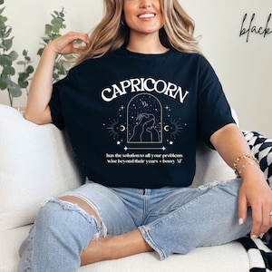 Puede incluir: Camiseta negra con un dise&ntilde;o gr&aacute;fico blanco que representa el signo del zod&iacute;aco Capricornio. El gr&aacute;fico incluye el texto "CAPRICORN" y la frase "has the solution to all your problems wise beyond their years - bossy AF".