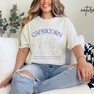 Puede incluir: Una camiseta beige claro con un gr&aacute;fico morado del signo astrol&oacute;gico Capricornio. El gr&aacute;fico incluye el s&iacute;mbolo de Capricornio, una luna creciente y estrellas. El texto "CAPRICORN" est&aacute; impreso en morado sobre el gr&aacute;fico. El texto "has the solution to all your problems wise beyond their years + bossy AF" est&aacute; impreso en negro debajo del gr&aacute;fico.