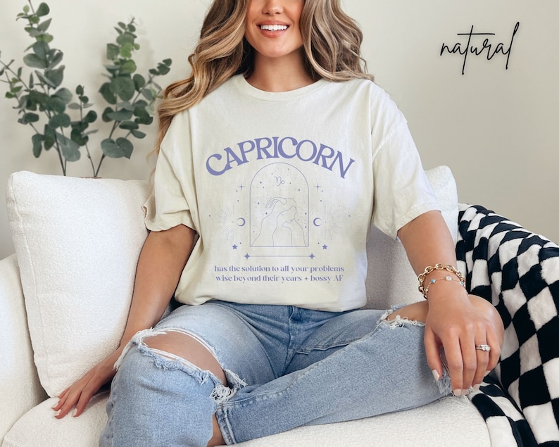 Puede incluir: Una camiseta beige claro con un gr&aacute;fico morado del signo astrol&oacute;gico Capricornio. El gr&aacute;fico incluye el s&iacute;mbolo de Capricornio, una luna creciente y estrellas. El texto "CAPRICORN" est&aacute; impreso en morado sobre el gr&aacute;fico. El texto "has the solution to all your problems wise beyond their years + bossy AF" est&aacute; impreso en negro debajo del gr&aacute;fico.
