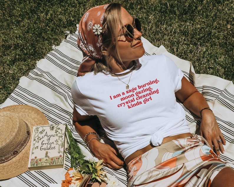 K&ouml;nnte beinhalten: Ein wei&szlig;es T-Shirt mit dem Text "I am a sage burning, moon gazing, crystal cleansing kinda girl" in roten Buchstaben. Das Shirt wird von einer Person getragen, die auf einer gestreiften Decke mit einem Buch mit dem Titel "Anne of Green Gables" und einem Blumenstrau&szlig; sitzt.