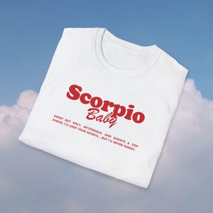 May include: White t-shirt with red text that reads "Scorpio Baby" and a description below: "Sweet but spicy, mysterious, and always a step ahead. I'll keep your secrets... but I'll never forget."