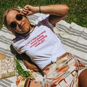 K&ouml;nnte beinhalten: Ein wei&szlig;es T-Shirt mit dem Text "I am a sage burning, moon gazing, crystal cleansing kinda girl" in Rot auf der Vorderseite bedruckt. Das Shirt wird von einer Person getragen, die auf einer gestreiften Decke liegt.