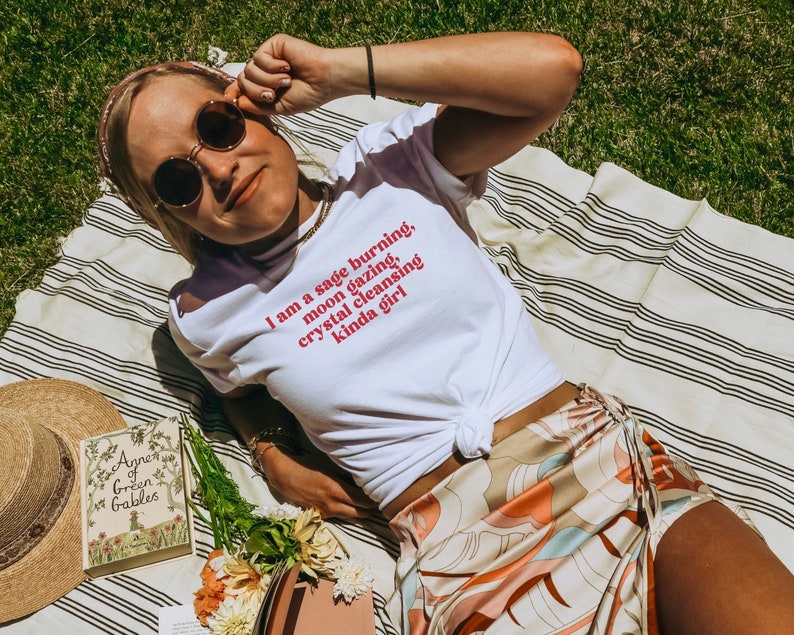 K&ouml;nnte beinhalten: Ein wei&szlig;es T-Shirt mit dem Text "I am a sage burning, moon gazing, crystal cleansing kinda girl" in Rot auf der Vorderseite bedruckt. Das Shirt wird von einer Person getragen, die auf einer gestreiften Decke liegt.