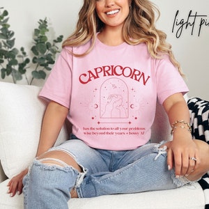 Puede incluir: Camiseta rosa claro con un gr&aacute;fico rojo del signo zodiacal Capricornio. El gr&aacute;fico incluye el texto "CAPRICORN", el s&iacute;mbolo de Capricornio y el texto "has the solution to all your problems wise beyond their years + bossy AF".
