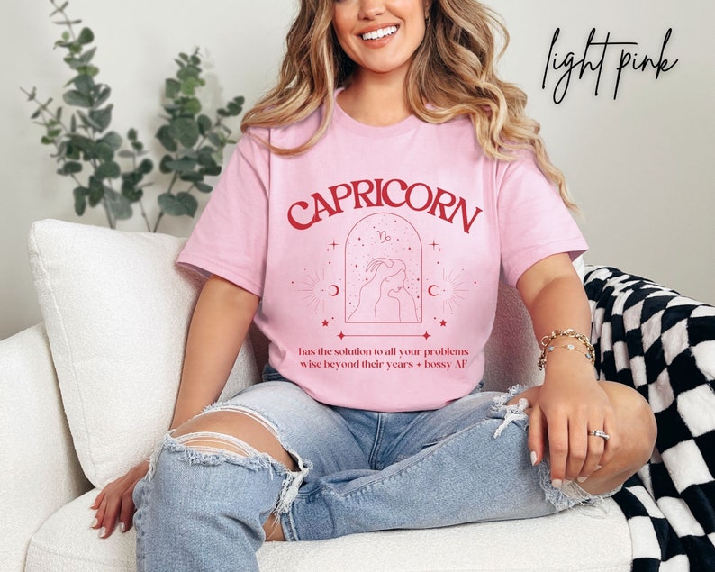 Puede incluir: Camiseta rosa claro con un gr&aacute;fico rojo del signo zodiacal Capricornio. El gr&aacute;fico incluye el texto "CAPRICORN", el s&iacute;mbolo de Capricornio y el texto "has the solution to all your problems wise beyond their years + bossy AF".