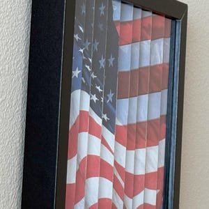 Pu&ograve; includere: Opera d'arte incorniciata con la bandiera americana. Il design della bandiera &egrave; visibile attraverso fessure verticali, creando un effetto visivo unico. La cornice &egrave; nera e la bandiera mostra le tradizionali strisce rosse e bianche e il campo blu con stelle bianche.