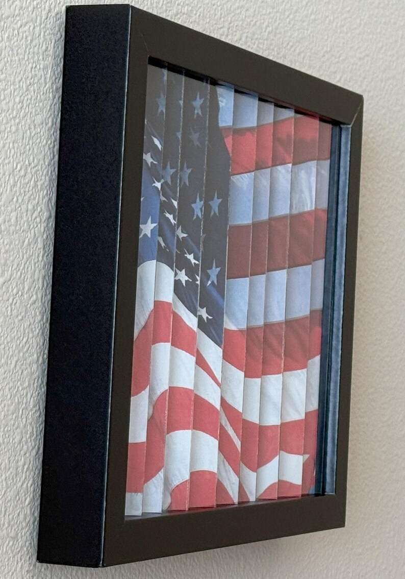 Pu&ograve; includere: Opera d'arte incorniciata con la bandiera americana. Il design della bandiera &egrave; visibile attraverso fessure verticali, creando un effetto visivo unico. La cornice &egrave; nera e la bandiera mostra le tradizionali strisce rosse e bianche e il campo blu con stelle bianche.