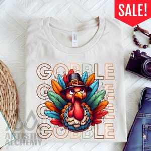 May include: A cream-colored t-shirt featuring a graphic of a turkey wearing a black pilgrim hat. The turkey has colorful red, orange, blue, and green feathers. The text GOBBLE is printed in large, outlined letters behind the turkey illustration.