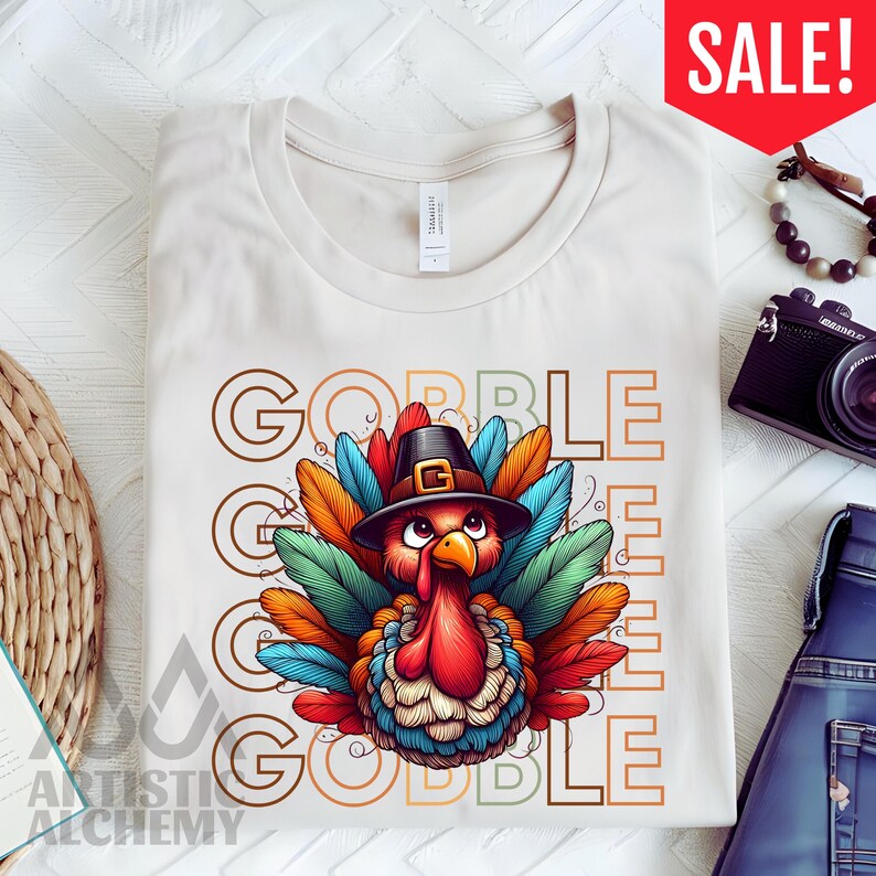 May include: A cream-colored t-shirt featuring a graphic of a turkey wearing a black pilgrim hat. The turkey has colorful red, orange, blue, and green feathers. The text GOBBLE is printed in large, outlined letters behind the turkey illustration.