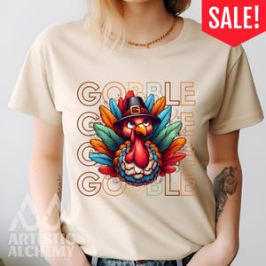 May include: A beige short-sleeve t-shirt featuring a central graphic of a turkey wearing a black pilgrim hat. The turkey has colorful red, blue, and orange feathers. The text GOBBLE GOBBLE GOBBLE is printed in a repeating outline font behind the turkey.