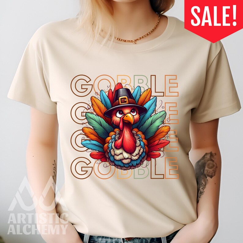 May include: A beige short-sleeve t-shirt featuring a central graphic of a turkey wearing a black pilgrim hat. The turkey has colorful red, blue, and orange feathers. The text GOBBLE GOBBLE GOBBLE is printed in a repeating outline font behind the turkey.