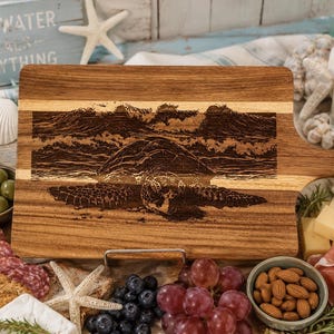May include: Wooden cutting board with a handle, featuring an engraved image of a sea turtle and ocean waves. The board is made of light and dark wood tones. The text "SALTWATER HEALS EVERYTHING" is visible in the background.