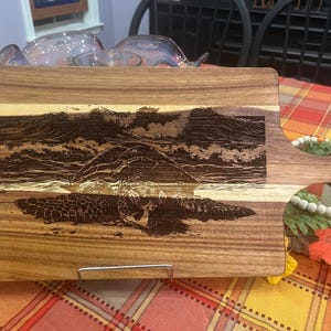 Op de afbeelding: Houten snijplank met een handvat, met een gegraveerd ontwerp van een zeeschildpad die voor een golf zwemt. De plank is rechthoekig met een handvat en een metalen standaard.