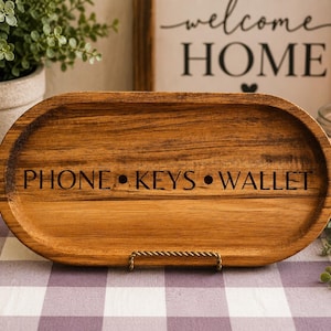 Puede incluir: Una bandeja ovalada de madera con las palabras "PHONE • KEYS • WALLET" grabadas en negro. La bandeja es de madera marrón claro y se apoya en un soporte de metal dorado. El fondo incluye un letrero de "welcome HOME" y una vela encendida.