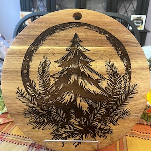 May include: Round wooden cutting board with a dark etched design of a pine tree surrounded by pine needles. The board has a hole for hanging and is displayed on a metal stand. The wood grain is visible, and the design is in a circular frame.