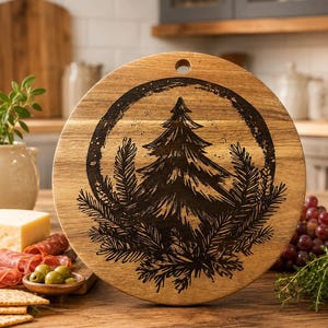 May include: A round wooden serving board with a dark etched design of a Christmas tree and pine branches. The board has a hole for hanging and is surrounded by food items like cheese, crackers, olives, and grapes. The board is on a wooden surface.