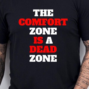 May include: Black crew neck t-shirt with the text "THE COMFORT ZONE IS A DEAD ZONE" in white and red lettering. The shirt is worn by a person with tattoos on their arm.
