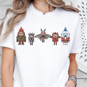 Op de afbeelding: Wit T-shirt met een rij cartoonfiguren: een kabouter, een gehoornd wezen, een vleermuisachtig figuur, een duivel en de kerstman. De figuren zijn in verschillende kleuren en staan naast elkaar. Het T-shirt wordt gedragen.