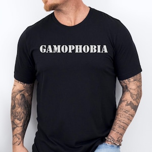 Puede incluir: Camiseta negra de cuello redondo con la palabra "GAMOPHOBIA" impresa en letras blancas. La camiseta la lleva una persona con tatuajes en los brazos y se combina con unos vaqueros azules.