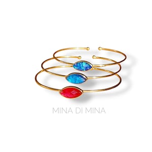 Op de afbeelding: Drie goudkleurige manchetarmbanden, elk met een marquise-vormige edelsteen. De edelstenen zijn blauw, blauwgroen en rood. De armbanden zijn op een witte achtergrond gestapeld. De tekst "MINA DI MINA" staat eronder.