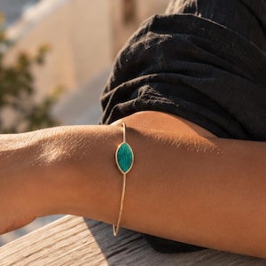 May include: A thin gold-toned metal bangle bracelet featuring a central marquise-shaped inlay of green malachite with distinct dark banding patterns. The bracelet has a simple, open-ended design that rests securely around the wrist.