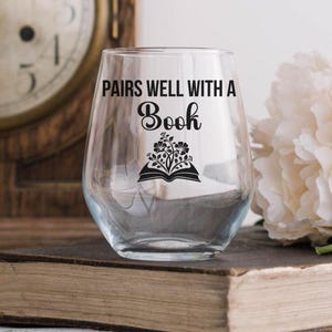 May include: A clear stemless wine glass with the words "PAIRS WELL WITH A Book" in black script. Below the text is a black graphic of an open book with flowers. The glass is on a thick, old book.